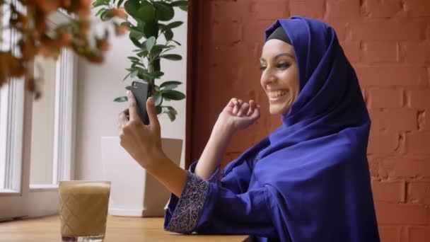 Young muslim woman in hijab having video chat through cell phone, smiling, sitting in modern cafe - 映像、動画