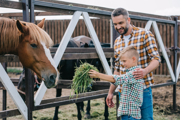 happy father looking at little son feeding horse at farm - Photo, Image