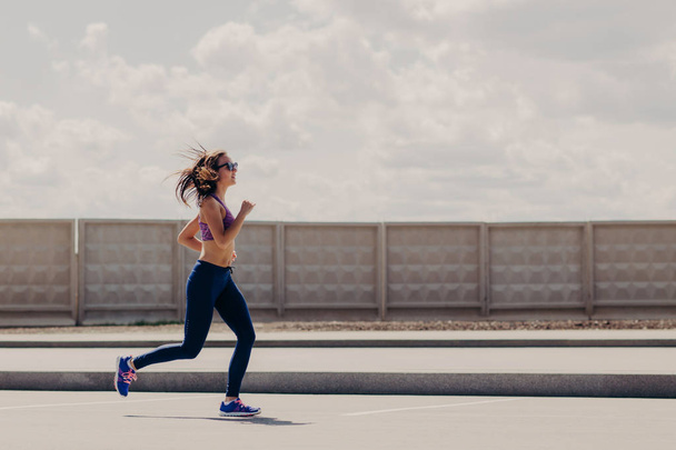 Horizontal shot of sportive woman joggs in open air, enjoys sport activities, wears sport clothes, concentrated into distance, has running workout. Female runner motivates yuo to go in for sport - Foto, Bild