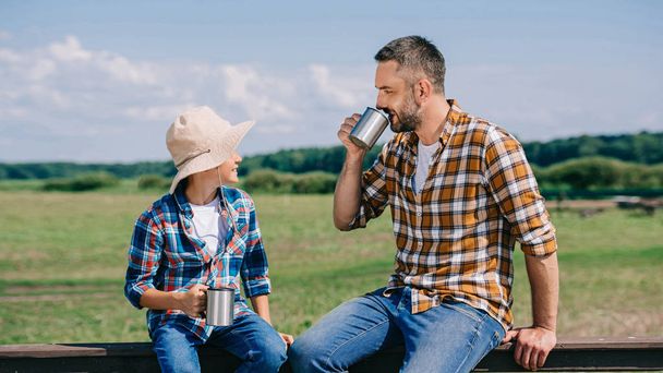 feliz padre e hijo sentado en la valla y beber de copas de metal en la granja
 - Foto, Imagen