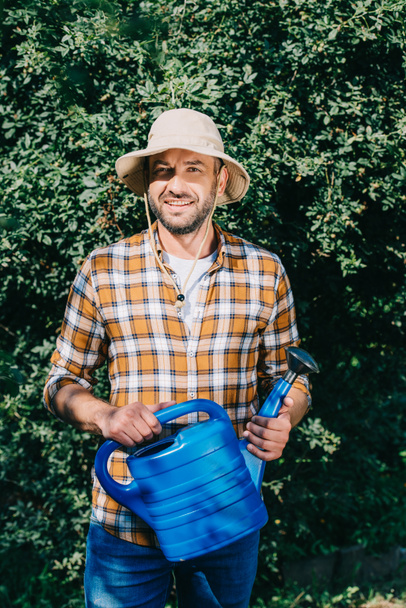 handsome male farmer in checkered shirt holding watering can and smiling at camera outdoors - Zdjęcie, obraz