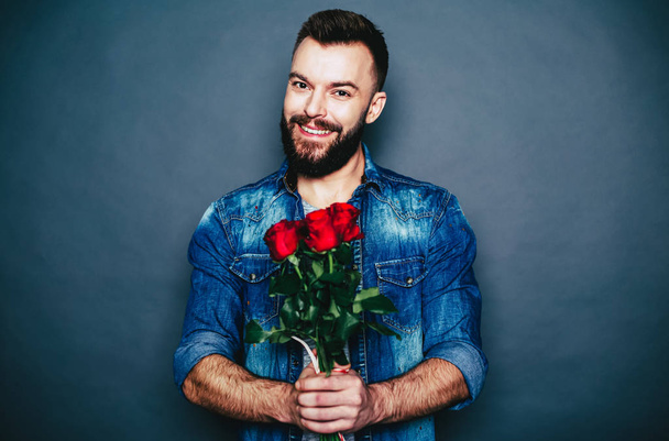 Young smiling man holding red roses over grey background - Fotoğraf, Görsel