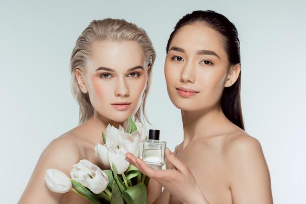beautiful naked multicultural women posing with tulips and bottle of perfume, isolated on grey - Foto, afbeelding