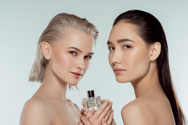 naked multiethnic girls posing with bottle of perfume, isolated on grey - Photo, Image
