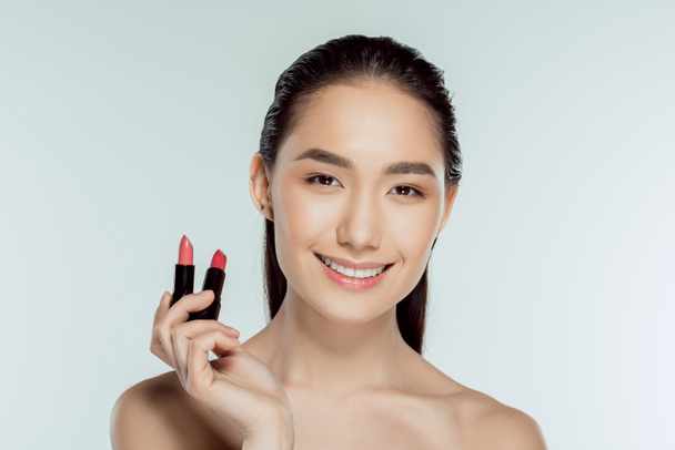 attractive smiling asian woman holding two lipsticks, isolated on grey - Photo, Image