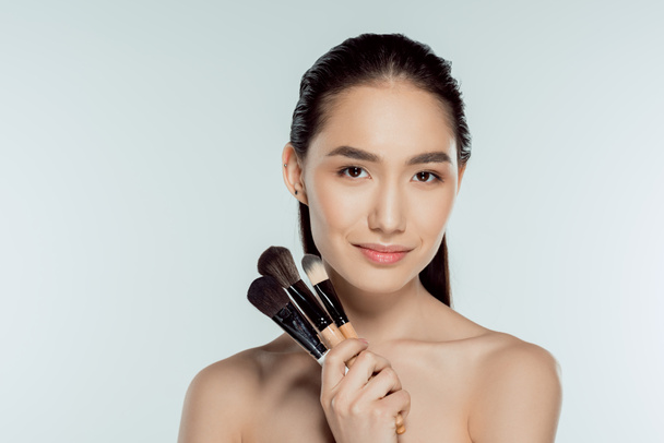 beautiful asian girl holding makeup brushes, isolated on grey - Фото, изображение