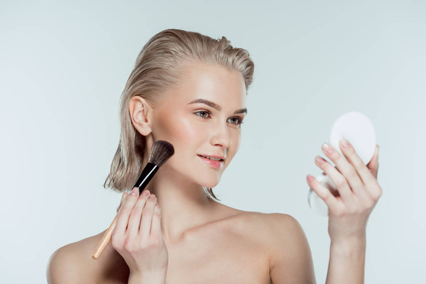 blonde woman applying compact powder with cosmetic brush, isolated on grey - Фото, изображение