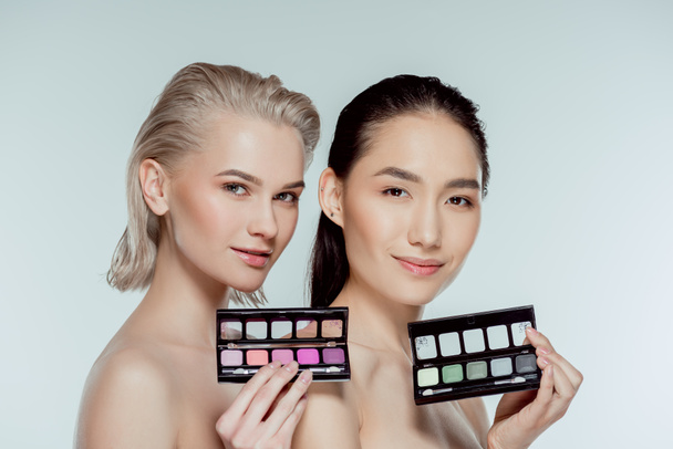 multiethnic naked women posing with palettes of eyeshadows, isolated on grey - Foto, Imagen