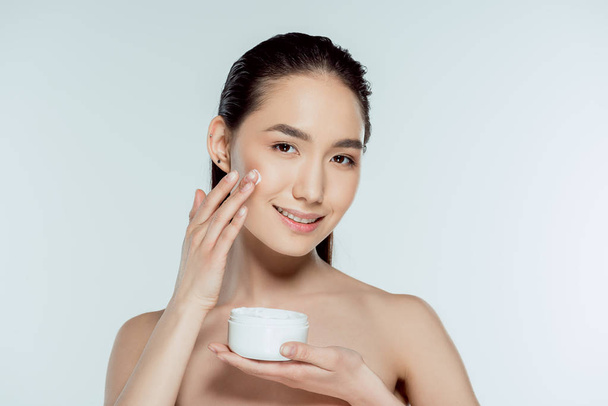 attractive asian woman applying face cream, isolated on grey - Фото, изображение