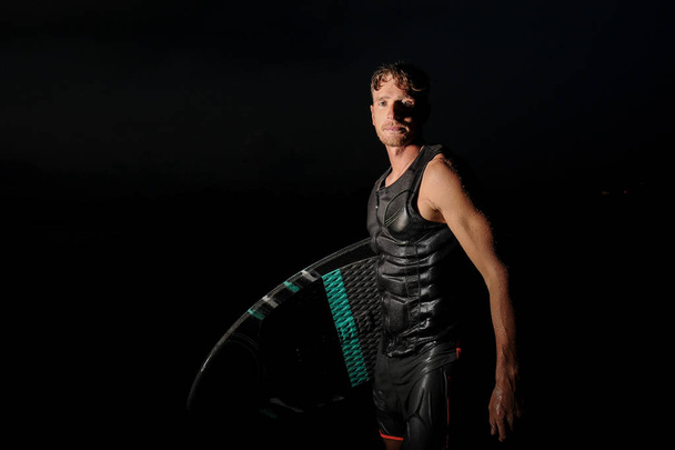 Handsome and muscular wakesurfer standing on the dark background of black and hot summer night - Foto, imagen