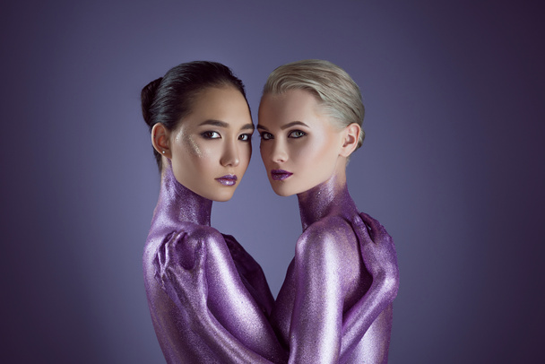 красивые мультикультурные девушки в ультрафиолетовом блеске, обнимающие друг друга, изолированные на фиолетовый
 - Фото, изображение