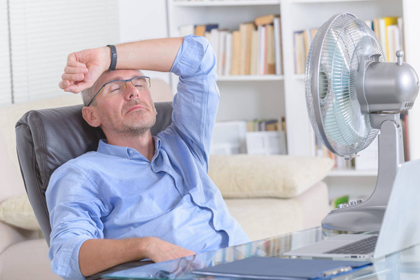 Чоловік страждає від спеки під час роботи в офісі і намагається охолодити вентилятор
 - Фото, зображення
