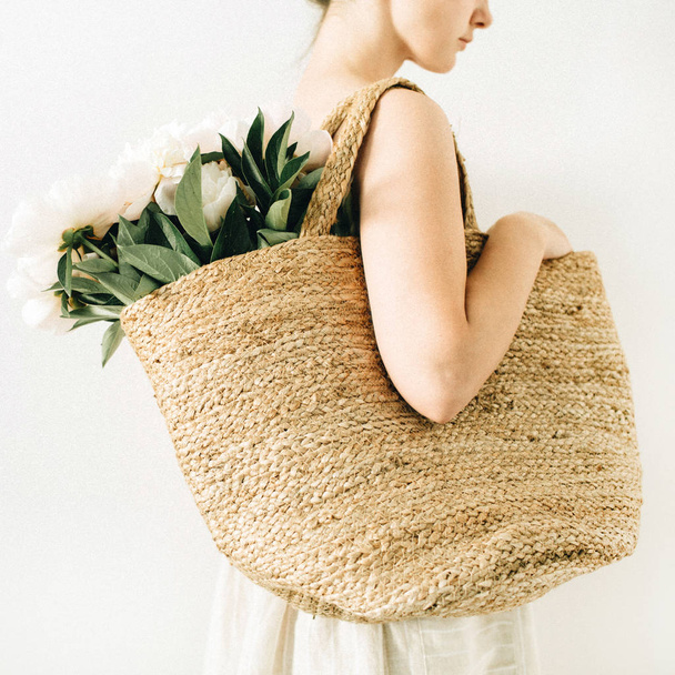 Jonge mooie vrouw met stro tas met witte pioen bloemen boeket op witte achtergrond. Zomer fashion concept. - Foto, afbeelding