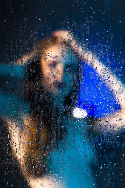 Silueta borrosa de una hermosa chica desnuda delgada de pie detrás de un cristal cubierto de gotas de agua. Foto artística y conceptual en tonos azules. Copiar espacio
. - Foto, Imagen