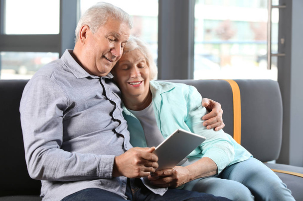 Счастливая пожилая пара с планшетным компьютером сидит дома на диване
 - Фото, изображение