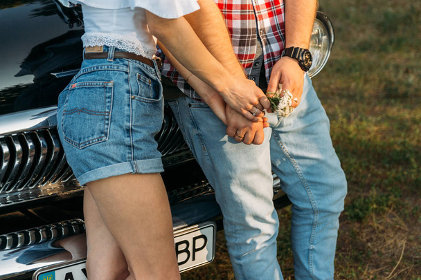 torso of a man and a woman holding hands, with a flower and with a clock on his hand. standing near the car.check shirt, shorts and jeans, white blouse - Foto, afbeelding