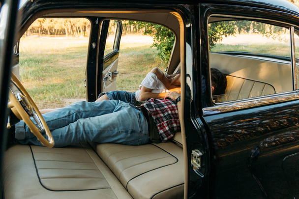 kissing people who are lying in the car and holding hands. look through the window in the car.side view.dressed in a plaid shirt, shorts and jeans, a white blouse. doors in the car are open, seats are spread out. black car with leather, white seats.d - Foto, afbeelding