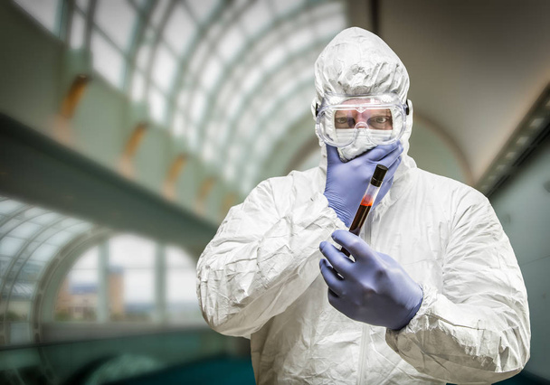 Mann in Schutzkleidung hält blutgefülltes Reagenzglas im Gebäude. - Foto, Bild
