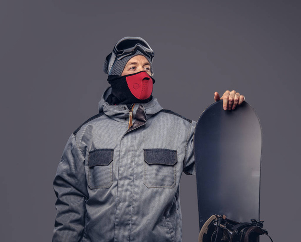 Πορτρέτο του ένας snowboarder ντυμένος με μια πλήρη προστατευτικό εξοπλισμό για extream snowboarding ποζάρει με snowboard σε ένα στούντιο. Απομονωμένοι σε ένα γκρίζο φόντο. - Φωτογραφία, εικόνα