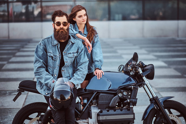 attraktives Hipster-Paar - bärtiger brutaler Mann mit Sonnenbrille und Jeansjacke sitzt auf einem Retro-Motorrad und sein junges sinnliches Mädchen steht daneben und posiert vor einem Wolkenkratzer. - Foto, Bild
