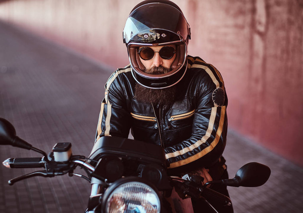 Close-up portrait of a brutal bearded biker in helmet and sunglasses dressed in a black leather jacket sitting on a retro motorcycle with included headlight. - Fotó, kép