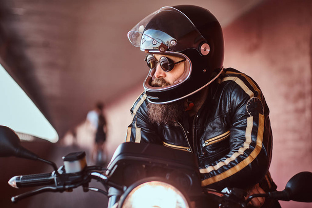 Close-up portrait of a brutal bearded biker in helmet and sunglasses dressed in a black leather jacket sitting on a retro motorcycle with included headlight. - Photo, Image