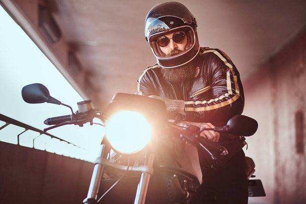 Retrato de cerca de un brutal motorista barbudo con casco y gafas de sol vestido con una chaqueta de cuero negro sentado en una motocicleta retro con faros incluidos
. - Foto, Imagen