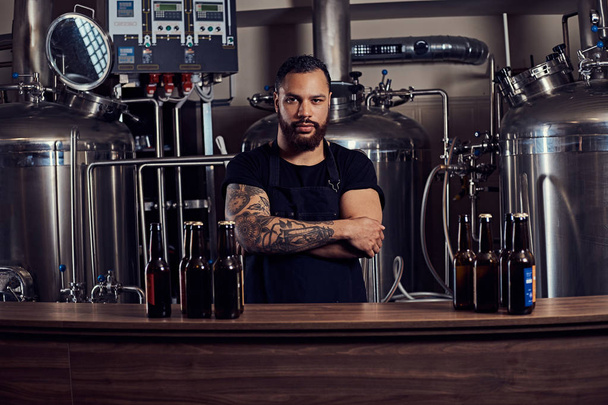 Portrait of a stylish bearded tattooed dark skinned male with crossed arms standing behind the counter in the brewery. - Photo, Image