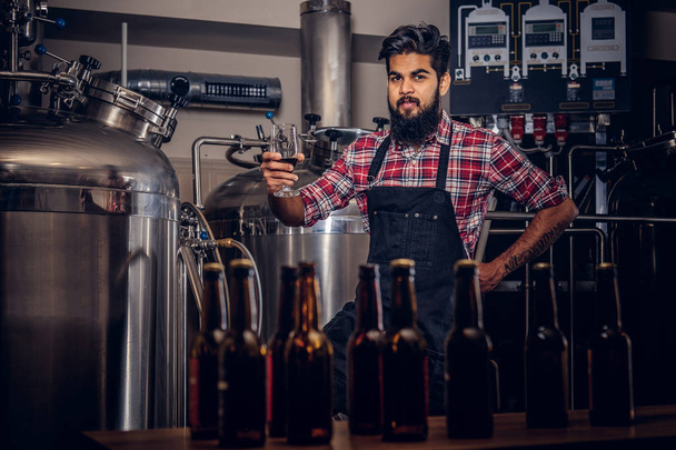 Μοντέρνο πλήρως γενειοφόρος ινδική άνθρωπος σε ένα πουκάμισο fleece και ποδιά κατέχει ένα ποτήρι μπίρα, στέκεται πίσω από τον πάγκο σε ένα ζυθοποιείο. - Φωτογραφία, εικόνα
