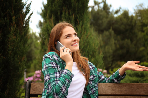 Młoda kobieta rozmawia przez telefon komórkowy siedząc na drewnianej ławce w parku - Zdjęcie, obraz