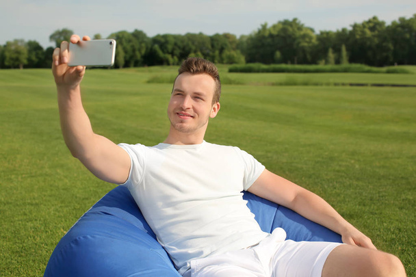 junger Mann macht Selfie, während er im Freien auf einem Bohnensackstuhl sitzt - Foto, Bild