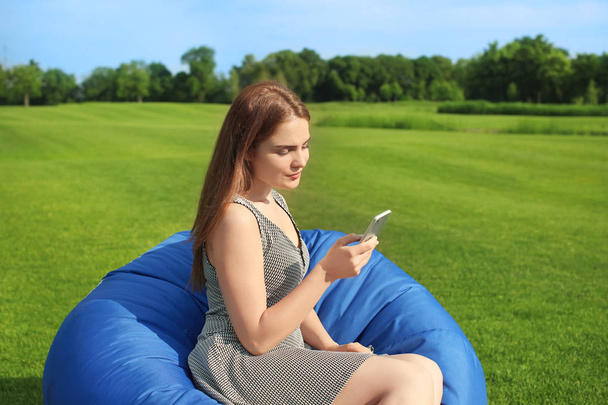 Молода жінка з мобільним телефоном відпочиває на стільці на відкритому повітрі
 - Фото, зображення
