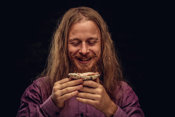 Close-up retrato de um feliz hipster ruiva macho com cabelos longos e barba cheia vestida com uma camisa roxa mantém um pequeno pires com chá. Isolado em fundo escuro texturizado
. - Foto, Imagem