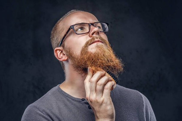 Крупный план портрета рыжеволосого бородатого мужчины в очках в серой футболке, заботящегося о своей бороде с помощью щетки для бороды. Изолированный на темном текстурированном фоне
. - Фото, изображение