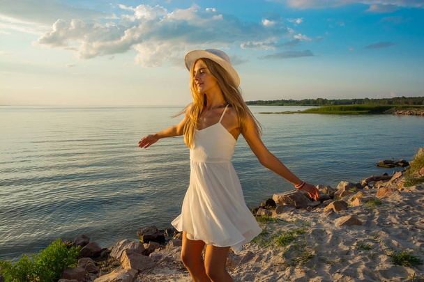 Szczęśliwy, piękną, młodą kobietę w biały lato sukienka z satyny i w kapeluszu wiry w tle morze o zachodzie słońca. - Zdjęcie, obraz