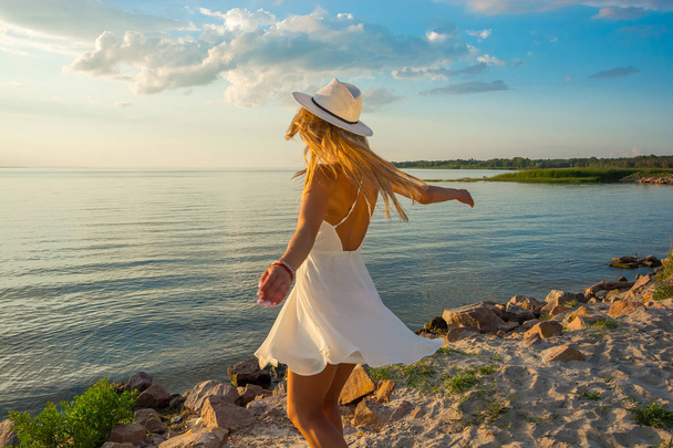 Красива щаслива молода жінка спина в білій літній атласній сукні і носить капелюхи на морському фоні на заході сонця
. - Фото, зображення