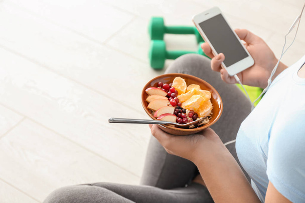 Sporty young woman with bowl of tasty oatmeal and mobile phone at home - Photo, image