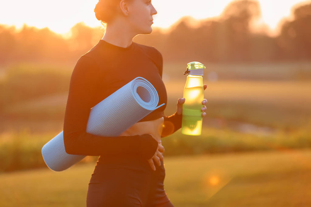 Mooie jonge vrouw met fles water en yoga mat buiten in de ochtend - Foto, afbeelding