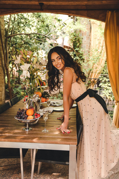 fashion outdoor photo of beautiful young woman with dark hair in elegant dress posing in lounge zone of a hotel. plate with fruits and glass of champagne on background - 写真・画像