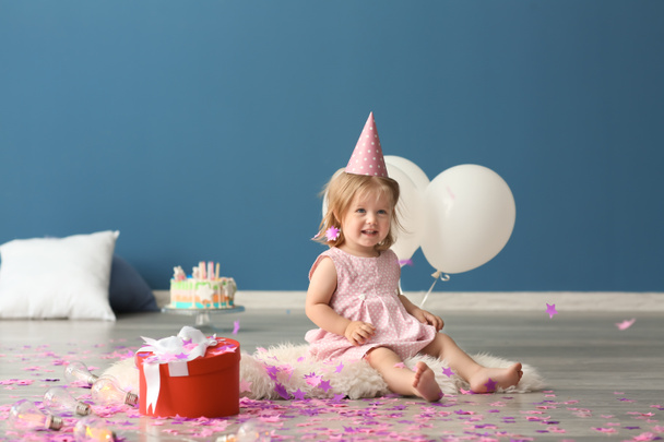 niedliches kleines Mädchen in Geburtstagskappe mit Geschenkbox sitzt auf pelzigen Teppich drinnen - Foto, Bild