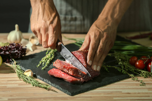 Mann schneidet rohes Fleisch auf Schieferteller in Küche - Foto, Bild