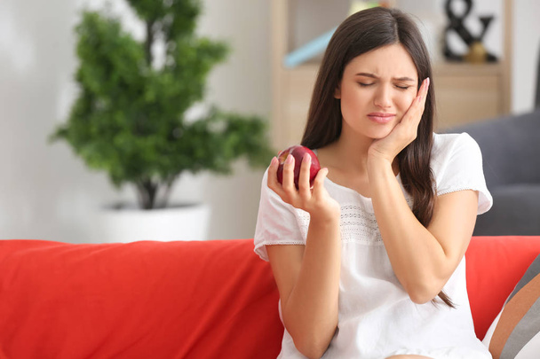 Young woman with sensitive teeth and apple on sofa at home - Photo, Image