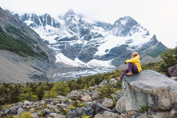 A girl in a yellow jacket and tracking clothes is sitting on the rocks in the background of the high snow-capped mountains - Photo, Image