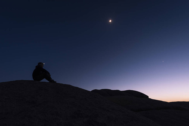 Silhouette of hiker sitting on rock formations with a headlamp lit watching the sunset and the purple and blue sky behind him with stars above in Joshua Tree National Park, California - Photo, Image