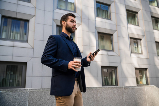 Jistý mladý businessmanin modrý oblek, pomocí mobilního telefonu ve městě. podnikatel pije kávu a textových zpráv venkovní úřad před budovy. - Fotografie, Obrázek