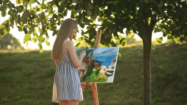 Hermosa chica de pie en el parque y dibuja un cuadro usando una paleta con pinturas y una espátula
. - Metraje, vídeo