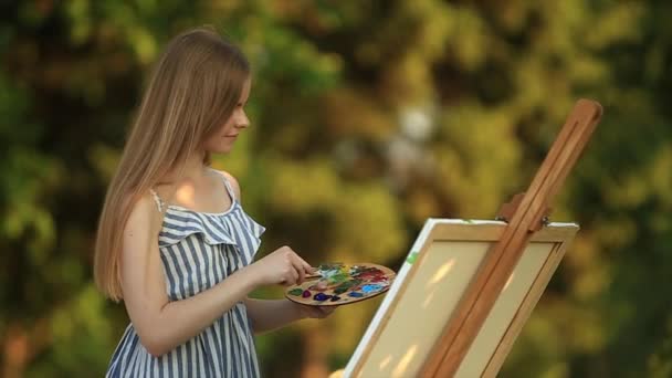 Όμορφο κορίτσι στέκεται στο πάρκο και τραβάει μια εικόνα χρησιμοποιώντας μια παλέτα με χρώματα και μια σπάτουλα. - Πλάνα, βίντεο