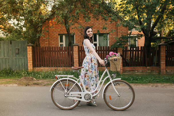 Smiling brunnete happy girl in a white-flowers dress with pink peones in the busket and vintage bicycle. Lovely house is on the background - Fotografie, Obrázek