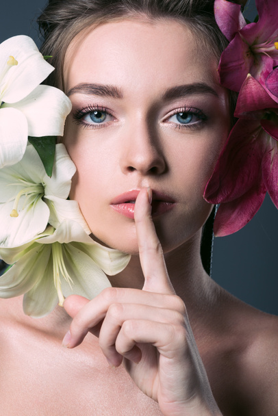 close-up portrait of beautiful young woman surrounded with lilium flowers showing silence gesture - Fotoğraf, Görsel