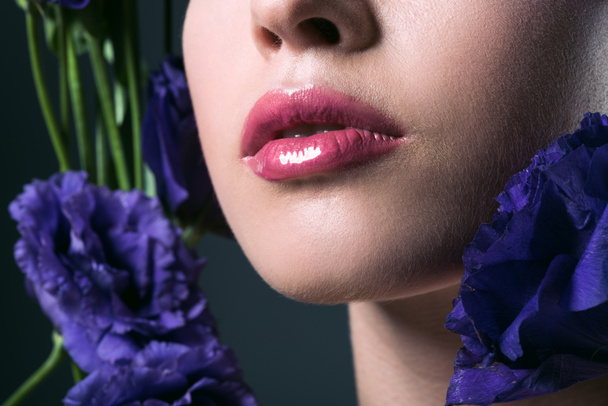 partial portrait of beautiful young woman with pink lips and purple eustoma flowers on grey - Foto, afbeelding